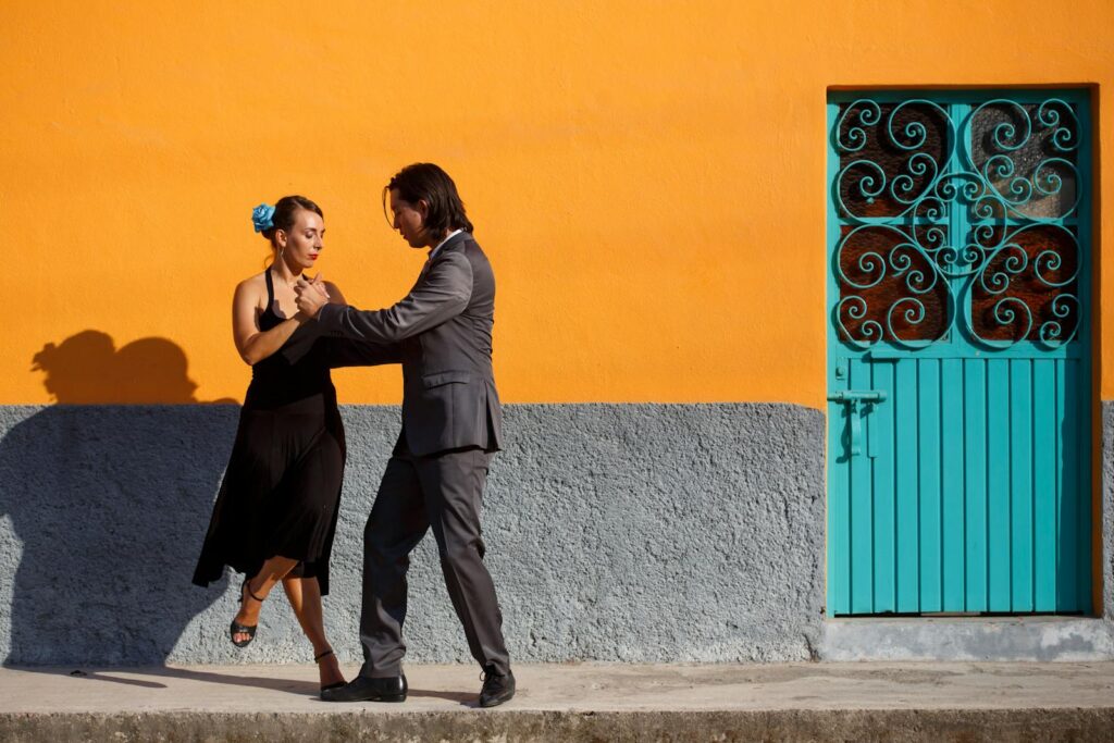 A couple performing a tango dance in front of a striking orange wall with a decorative turquoise door.
